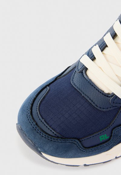 Sapatilha azul escura com uma mistura de couro liso e tecido texturizado. Os destaques incluem atacadores creme e um detalhe em verde no logótipo.