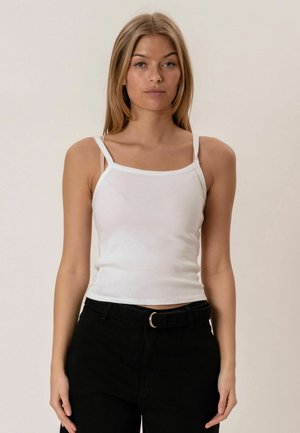 Joven mujer con cabello largo y rubio, vestida con una camiseta blanca sin mangas y acanalada, y pantalones negros con cinturón, de pie contra un fondo liso.