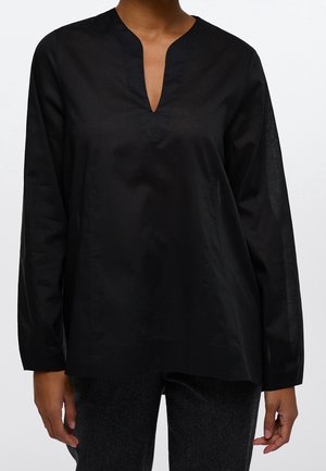 Femme portant un chemisier noir à manches longues avec un col en V et une coupe décontractée, ainsi qu'un pantalon gris foncé, debout devant un fond blanc uni.