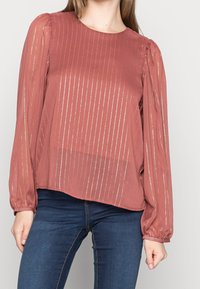 Blusa color óxido con mangas largas y voluminosas, con rayas verticales metálicas y cuello redondo; fabricada en una tela ligera y texturizada.