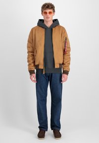 Chaqueta bomber de ante marrón con puños acanalados, sudadera gris, jeans de mezclilla azul y zapatos marrones. Las gafas de sol naranjas complementan el conjunto.