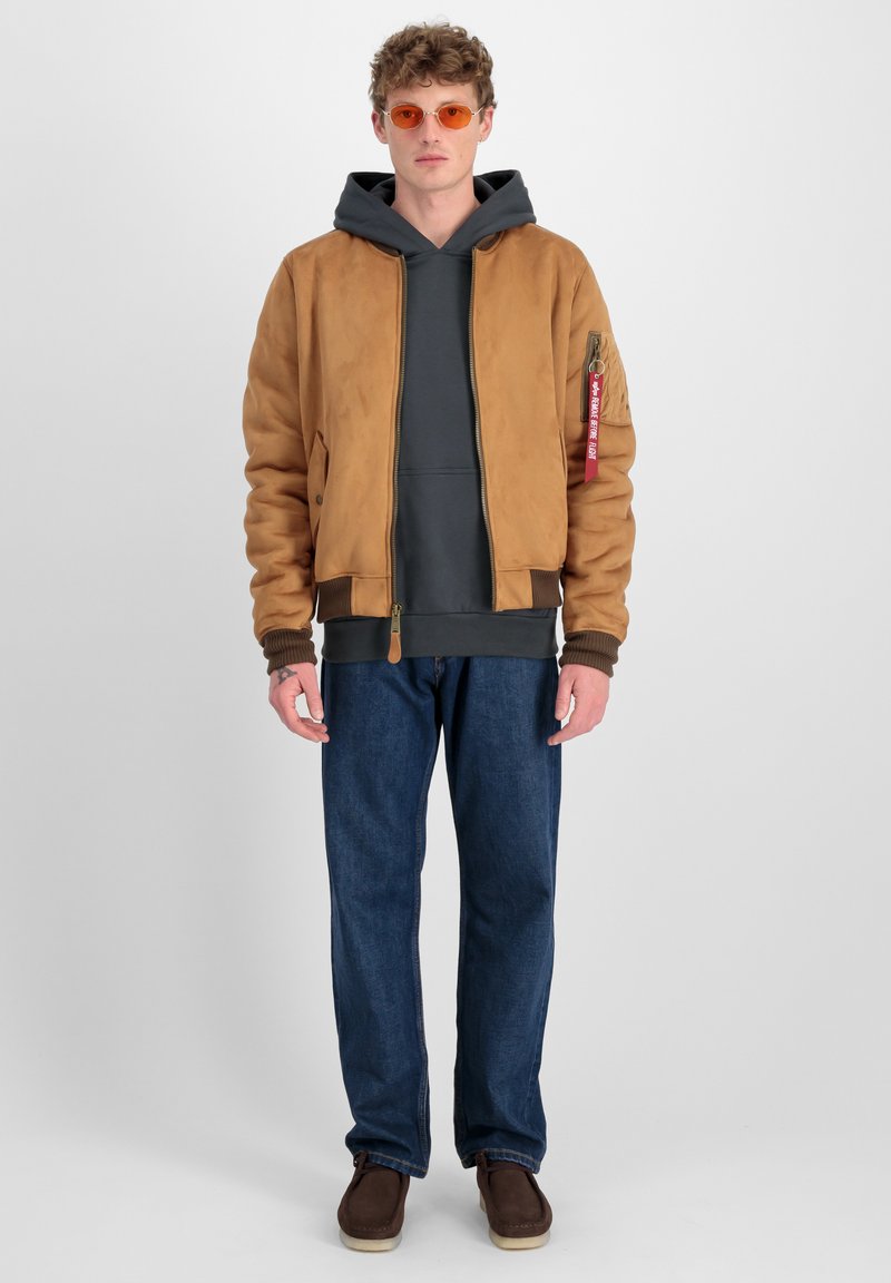 Chaqueta bomber de ante marrón con puños acanalados, sudadera gris, jeans de mezclilla azul y zapatos marrones. Las gafas de sol naranjas complementan el conjunto.