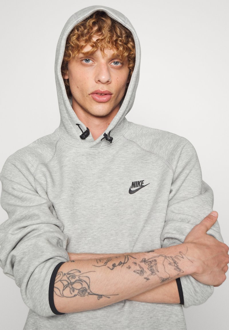 Sweat-shirt gris avec un logo Nike noir, capuche à cordon, poignets côtelés et coupe décontractée. Tatouages visibles sur les avant-bras, texture de tissu lisse.