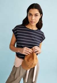 Navy striped short-sleeve shirt made of cotton, paired with beige trousers and a brown cap held in hands. Background is light blue.