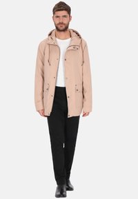 Beige Kapuzenjacke mit Kordelzug, Frontknopf- und Reißverschluss, zwei Fronttaschen, getragen über einem weißen Hemd und schwarzen Hosen. Schwarze Stiefel.