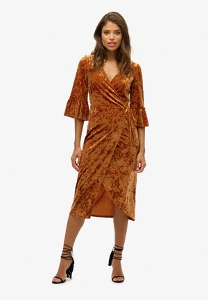 Vestido envolvente de terciopelo bronce metálico con mangas acampanadas de tres cuartos, dobladillo hasta la rodilla y detalle de lazo lateral. Combinado con tacones negros con tiras.