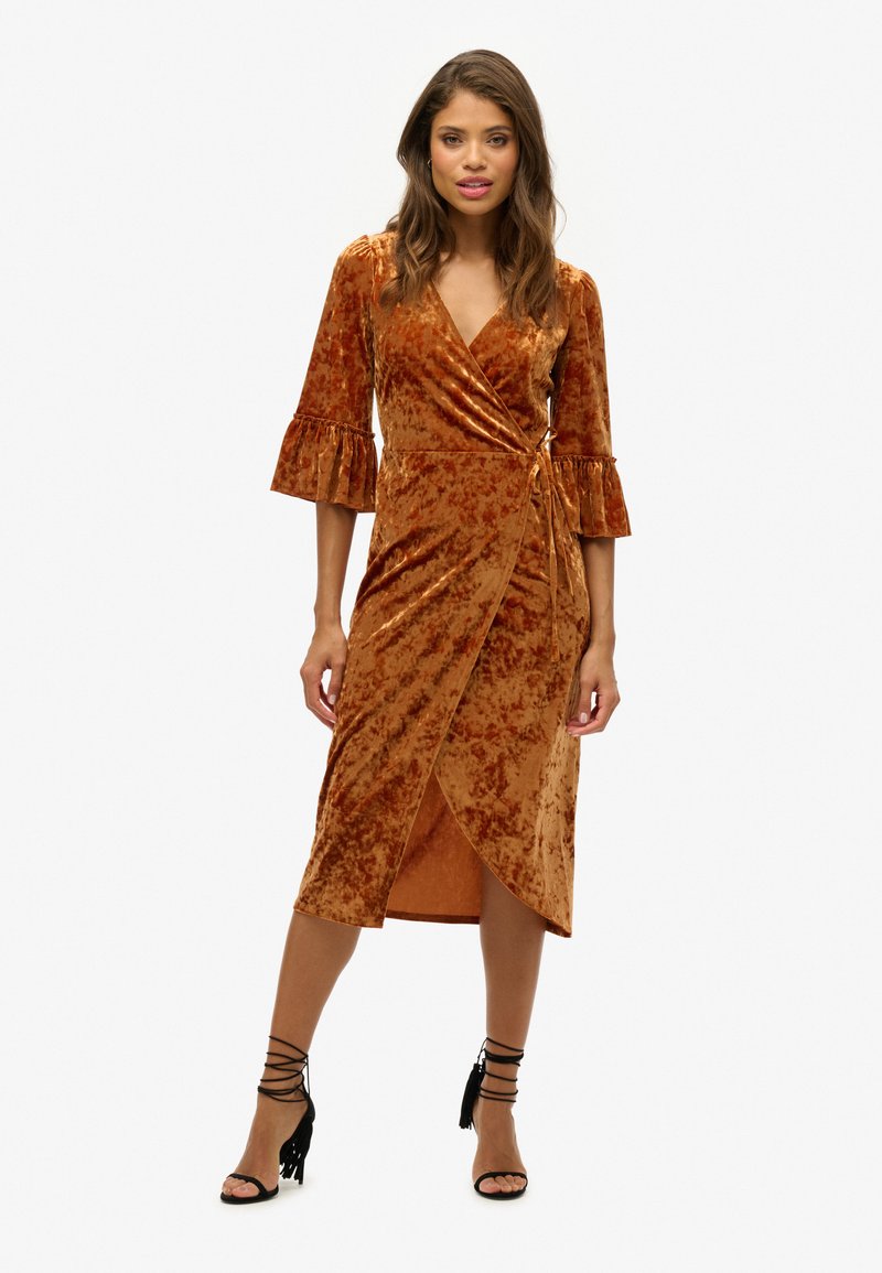 Robe enveloppante en velours bronze métallique avec des manches ruffl ées trois-quarts, une longueur mi-genou et un détail de nouage sur le côté. Associée à des talons noirs à lanières.