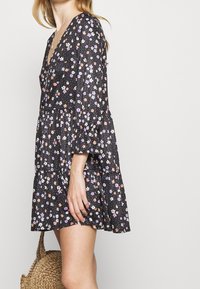 Robe florale noire avec un décolletée en V plongeant, des manches à cloche, un design en niveaux et un motif de petites fleurs violettes, roses et blanches.
