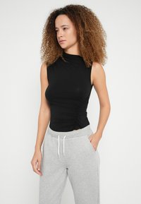 Svart ermeløs topp med høy krage, med rynkedetalj på den ene siden, kombinert med lysegrå joggebukser og hvite snorer.