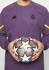 Sudadera morada con acentos amarillos, que presenta un logo negro. La persona sostiene un balón de fútbol con un diseño en negro, blanco y detalles naranjas.