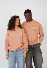 Two models wear oversized, peach-colored sweatshirts with a small printed design on the front. The backgrounds are neutral. Models also wear jeans.