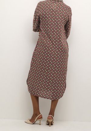 Vrouw met een knielange jurk met cirkelvormige bloemmotieven en beige slingback pumps met hoge hakken, staand tegen een effen lichte muur.