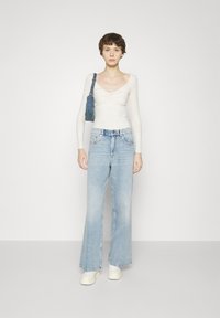 Top bianco a maniche lunghe con fronte attorcigliato, jeans a gamba larga blu chiaro e sneakers bianche, abbinati a una piccola borsa blu.