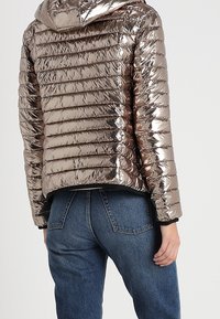 Giacca a vento bronzeo metallico con motivo a quadri orizzontali e cappuccio. Abbinata a jeans in denim blu, che mostrano uno stile casual e contemporaneo.