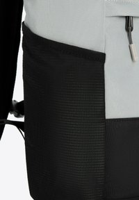 Sac à dos gris et noir avec une pochette en maille noire texturée, tissu lisse et fermeture éclair, comportant une petite étiquette de marque.