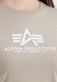 T-shirt en coton beige avec un logo graphique blanc présentant des lettres et des lignes stylisées. Encolure ronde et texture lisse.