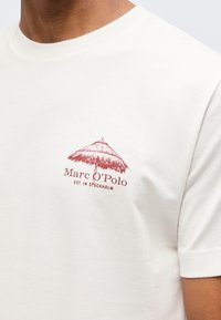Λευκό T-shirt με μικρό κόκκινο γραφικό μια ψάθινης ομπρέλας παραλίας και το κείμενο "Marc O'Polo EST. IN STOCKHOLM" στο στήθος.