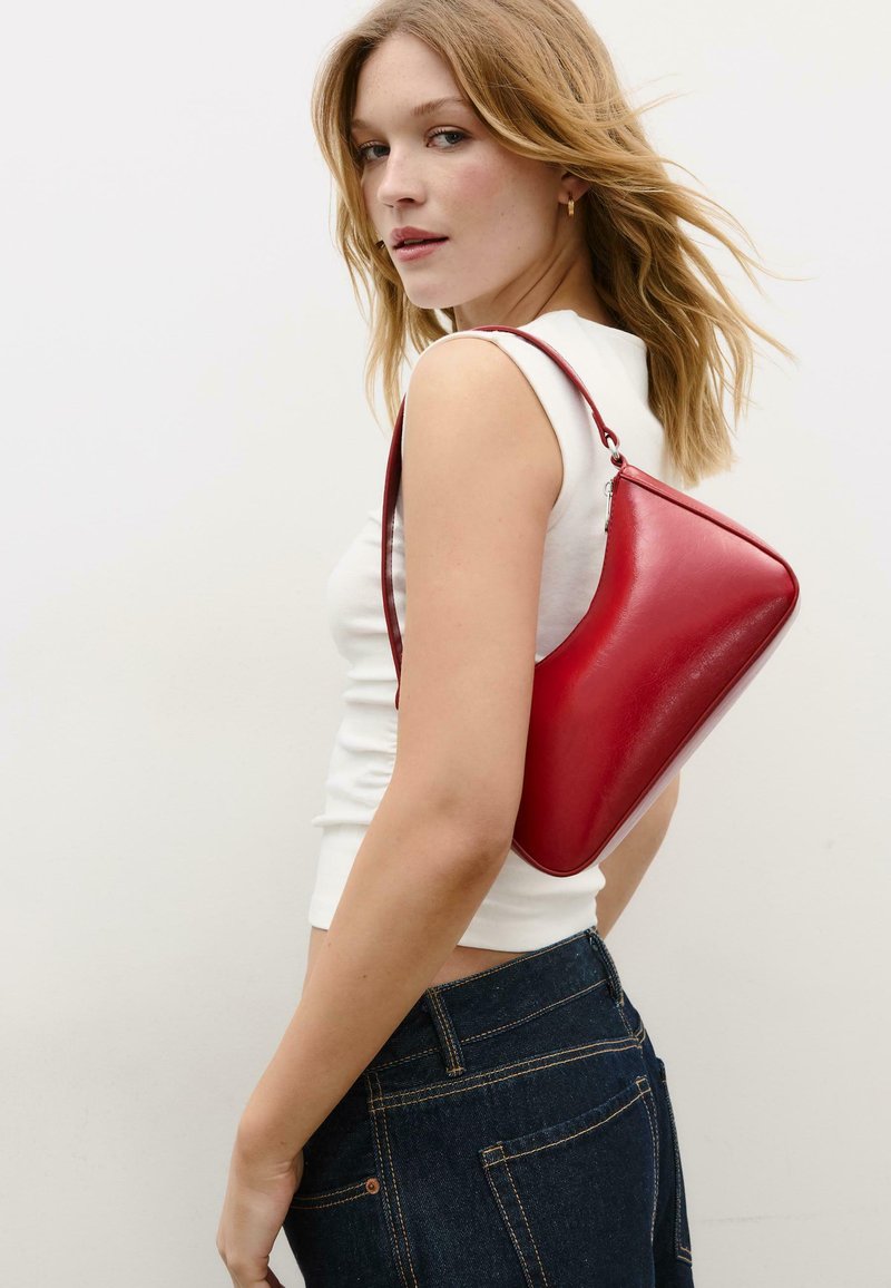 Jeune femme portant un haut blanc sans manches et un jean sombre, avec un sac à bandoulière en cuir rouge, regardant par-dessus son épaule.