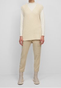 Pull sans manches beige en maille avec col en V, superposé à un col roulé blanc ajusté. Associé à un pantalon beige clair avec une poche latérale.