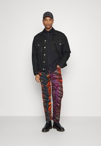 Giubbotto di jeans nero sopra una camicia nera, abbinato a pantaloni con strisce di tigre multicolori in arancione, viola e nero, e stivali Chelsea neri.