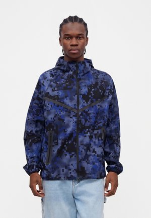 Ung mand med fletninger iført en blå og sort digital camouflettes jakke med lynlås og hætte samt lyseblå jeans.