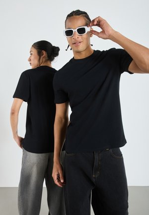 Svarta bomulls t-shirtar som bärs av två personer, en i mörka jeans och den andra i ljusgrå jeans. Vita solglasögon på mannen.