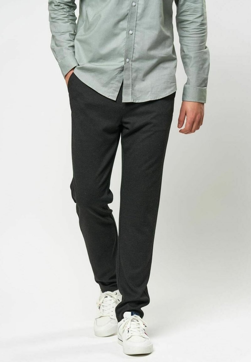 Mörkgrå joggingbyxor i mjukt tyg, med smal passform och elastisk midja, parat med en ljusgrön skjorta med knappar och vita sneakers.