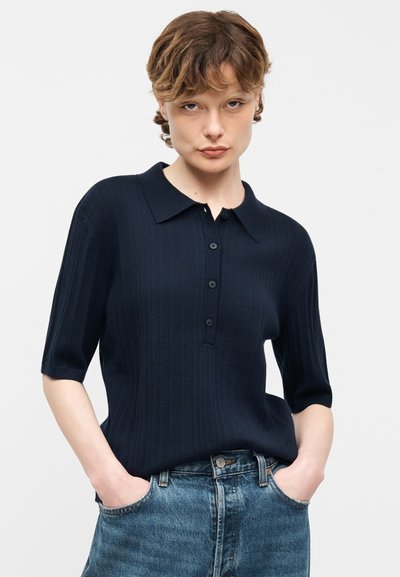 Νεαρός με κοντά καστανά μαλλιά, που φοράει μπλε σκούρο κοντομάνικο πόλο πουκάμισο με κουμπιά και μπλε τζιν, με τα χέρια στις τσέπες.