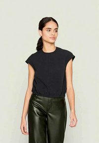 Mulher com cabelo escuro em um rabo de cavalo baixo, vestindo uma blusa preta sem mangas e calças de cabedal verde escuro, contra um fundo claro e liso.