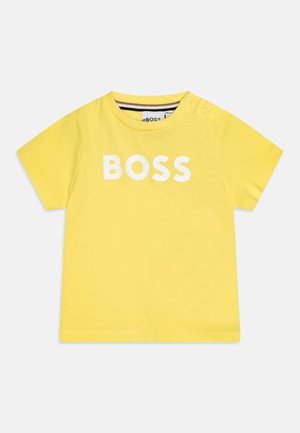 Gul kortærmet t-shirt til småbørn med hvid "BOSS" tekst foran og trykknapper nær halsudskæringen på en ensfarvet baggrund.