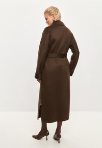 Manteau long en mélange de laine marron avec une taille ceinturée, un large col et des fentes latérales. Texture lisse et coupe ajustée.