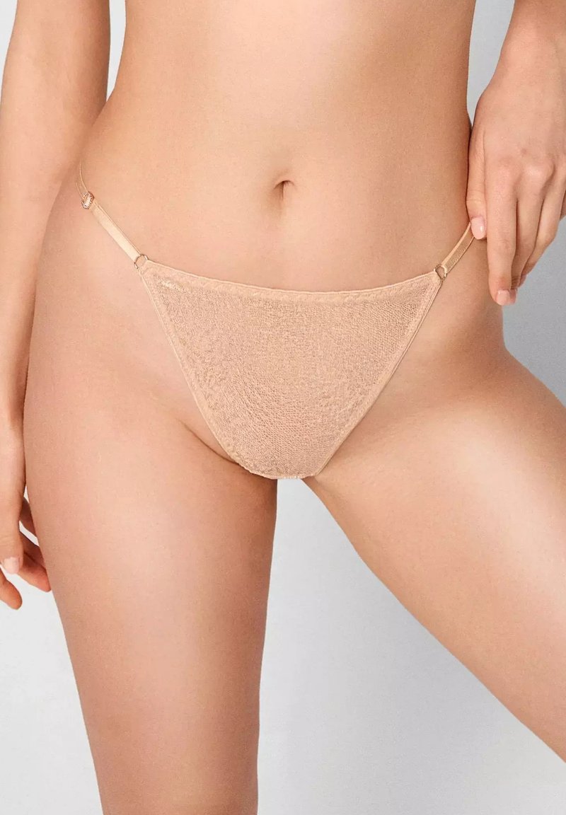 Beżowy string z miękką fakturą, trójkątnym przodem i cienkimi bocznymi paskami z metalowymi akcentami. Gładkie wykończenie podkreśla minimalistyczny design.