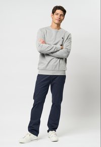 Hanorac gri cu manșete cu caneluri, pantaloni bleumarin și adidași albi. Design simplu, textură moale, fără modele. Modelul stă cu brațele încrucișate.