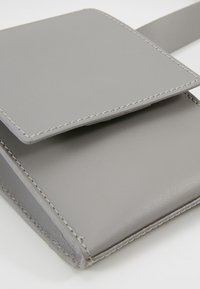 Šedá kožená peněženka s klopou, která se vyznačuje pečlivým šitím a hladkým povrchem. Kompaktní design s jednoduchou, moderní estetikou.