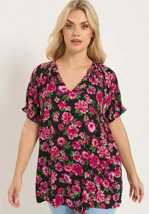 Blonde vrouw die een zwarte V-hals blouse draagt met een roze en paarse bloemmotief en korte mouwen, staand tegen een effen achtergrond.