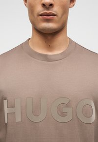 Κοντινή λήψη ενός ατόμου που φοράει μπεζ μπλούζα με στρογγυλή λαιμόκοψη και μεγάλα ανάγλυφα γράμματα που σχηματίζουν τη λέξη «HUGO» στο στήθος.