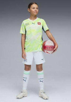 Jonge voetballer in fluorescerend groen en wit Zwitsers teamtenue, met een rood-witte bal met Zwitserse emblemen, staand op een grijze achtergrond.