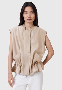 Gilet in suede beige con collo alto, chiusura a bottoni, pannello frontale a costine e vita a peplum elasticizzato. Dotato di tasche laterali e design senza maniche.