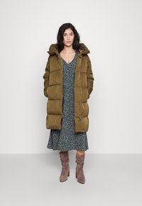 Olive žalia pūkų striukė su kapišonu, siuvinėtas dizainas, dėvima virš tamsiai mėlynos gėlių suknelės. Modelis taip pat dėvi pilkus plačius batus.