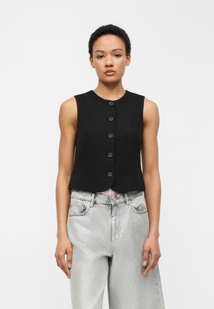 Frau trägt ärmellose schwarze Weste mit Knöpfen und hellgraue Jeans mit hohem Bund, steht vor schlichtem, hellem Hintergrund.
