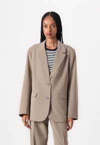 Beige skreddersydd blazer med en skarp klaff, enkelt knappelukking og påsydde lommer, kombinert med en svart og hvit stripete skjorte.