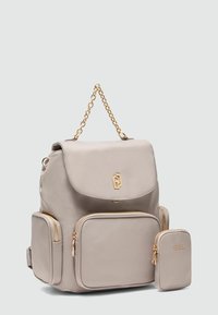 Sac à dos beige en matériau texturé avec une poignée en chaîne dorée, fermeture à rabat et deux poches avant zippées ; comprend un petit pochette assortie.