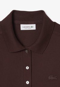 Polo shirt Lacoste marron foncé, avec un tissu texturé, une fermeture à trois boutons et un logo brodé discret sur la poitrine gauche.