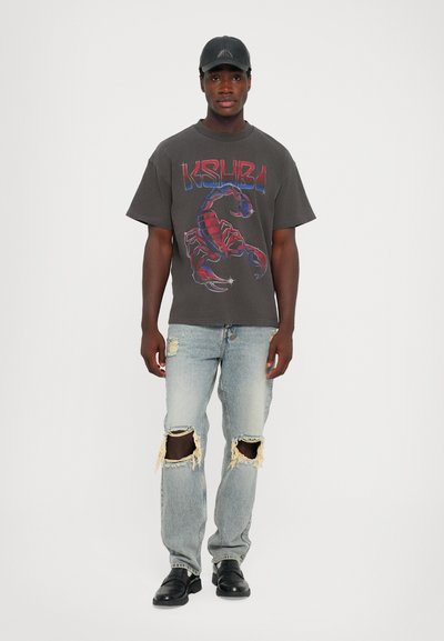 Tricou gri cu mânecă scurtă, având un grafic de scorpion roșu și albastru, asociat cu jeansi deschiși la culoare, cu tăieturi la genunchi, și pantofi negri.