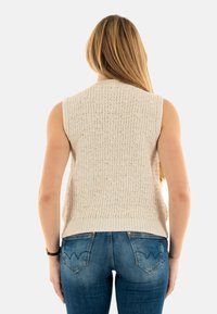 Jersey sin mangas beige con un patrón texturizado, cuello redondo y dobladillo de canalé, combinado con jeans de mezclilla azul que presentan un diseño de olas en los bolsillos.