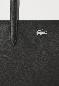 Sac fourre-tout en cuir noir texturé avec une longue sangle et un accent en logo de crocodile argenté. Présente une surface lisse et un design structuré.