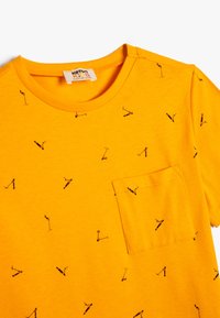 Gul T-shirt i bomull, med en ficka och ett upprepat mönster av svarta scootrar. Rund halsringning med förstärkt sömnad.