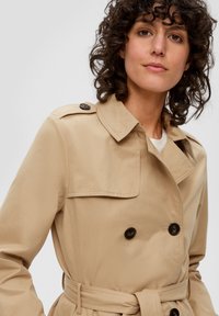 Manteau beige avec un design à double boutonnage, boutons noirs et taille ceinturée. Il présente un col cranté et une texture de tissu lisse.