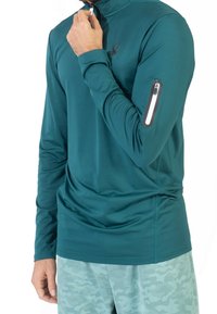 Camisa de manga larga color azul verdoso con un alto cuello, que cuenta con un bolsillo con cremallera en el brazo izquierdo y un logo de araña en el pecho. Textura suave.