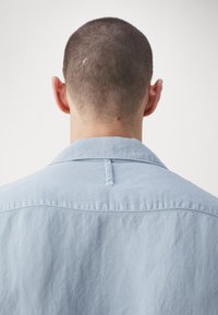 Camicia azzurra con finitura texture, dotata di colletto, dettaglio sul retro e asola per appenderla.